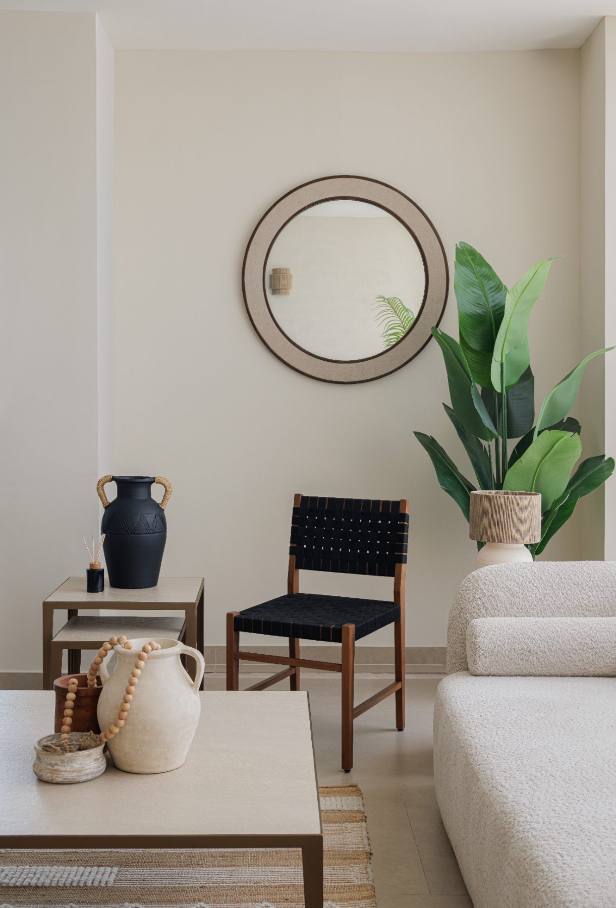 Round mirror above a black woven chair beside a leafy plant